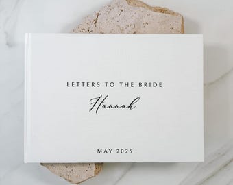 May include: A white rectangular book with a textured cover featuring the black printed text LETTERS TO THE BRIDE, the name Hannah in a cursive script, and the date MAY 2025 centered on the front cover in a clean, minimalist sans-serif font.