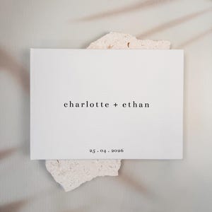 May include: A rectangular white book with a hard cover featuring the black printed text "charlotte + ethan" in the center and the date "25 . 04 . 2026" printed in black near the bottom edge of the front cover.