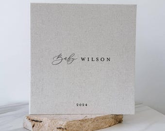 May include: A square, light beige, fabric-covered photo album or book with the text Baby WILSON printed in black, featuring a script font for Baby and a sans-serif font for WILSON, with the year 2024 printed in black at the bottom.