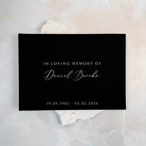 Op de afbeelding: Een rechthoekig zwart herinneringsboek met de in wit gedrukte tekst IN LOVING MEMORY OF, de naam Daniel Brooks in een schrijflettertype en de data 19.09.1982 - 03.02.2026 gecentreerd op de voorkant van het ingebonden boek.
