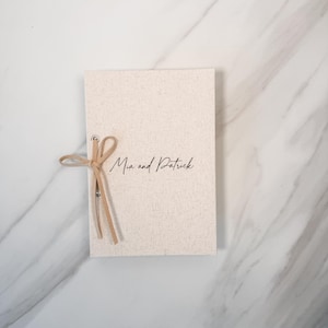 May include: A rectangular off-white fabric-covered notebook featuring the text "Mia and Patrick" in black script. The cover is secured with a light tan leather cord tied in a bow through two silver metal eyelets on the left side.