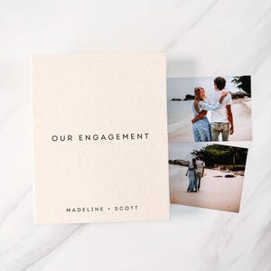 May include: A beige photo album with the words "OUR ENGAGEMENT" printed in black. Below the text are the names "MADELINE + SCOTT". Two photographs of a couple are next to the album. The photos show the couple on a beach.