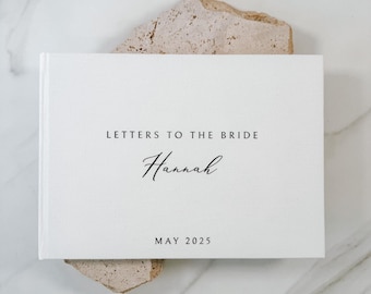 May include: A white rectangular book with a textured cover featuring the black printed text LETTERS TO THE BRIDE, the name Hannah in a cursive script, and the date MAY 2025. The book is positioned horizontally with a clean, simple design.