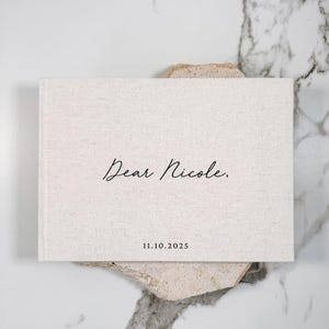 May include: A rectangular, off-white book with the text "Dear Nicole." and the date "11.10.2025" in black script. The book rests on a textured, light-colored stone against a marble background.