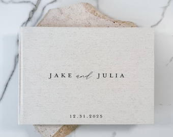 May include: A rectangular photo album with a light beige fabric cover. The front cover features the black printed text JAKE and JULIA in a mix of serif and script fonts, with the date 12.31.2025 printed in black below the names.