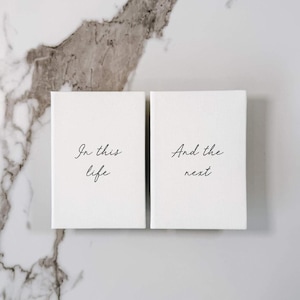 May include: Two rectangular white canvas panels featuring black cursive text. The left panel displays the words In this life, while the right panel displays the words And the next. Both panels have a clean, textured surface and sharp edges.