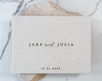 May include: A rectangular, light beige fabric-covered photo album featuring the black printed text JAKE and JULIA centered on the front cover, with the date 12.31.2025 printed in a smaller black font directly below the names.