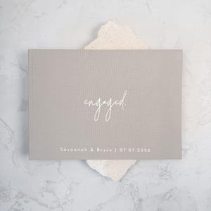May include: A rectangular light gray photo album with a fabric cover. The word "engaged." is printed in white script in the center, and the text "Savannah & Bryce | 07.07.2026" is printed in white sans-serif font at the bottom.