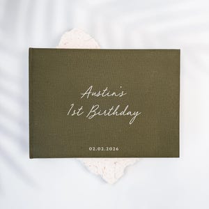 May include: A rectangular olive green photo album with a fabric-like texture. The cover features white text that reads Austin's 1st Birthday and the date 02.02.2026. The album is centered on a textured, off-white, irregular-shaped base.