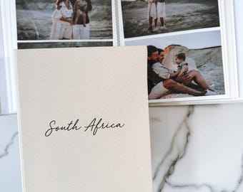 Könnte beinhalten: Ein beiges, stoffbezogenes Fotoalbum mit dem Text South Africa in schwarzer Schreibschrift und 2025 in schwarzem Druck auf dem Einband. Das geöffnete Album zeigt drei Fotos einer Familie in einer Sandlandschaft.