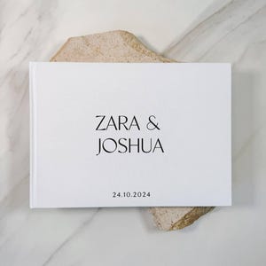 Könnte beinhalten: Ein weißes, rechteckiges Hardcover-Buch mit den Namen ZARA & JOSHUA, die in einer schwarzen Serifenschrift in der Mitte gedruckt sind. Unter den Namen ist das Datum 24.10.2024 in einer kleineren schwarzen Schriftart aufgedruckt.