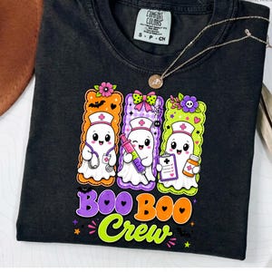 May include: A black t-shirt featuring a graphic of three white ghosts dressed as nurses, each inside a colorful rectangular border. The ghosts hold medical items including a stethoscope, a syringe, and a clipboard. The text reads BOO BOO Crew.