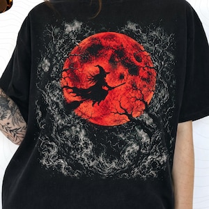 May include: A black t-shirt featuring a graphic print of a witch flying on a broomstick in front of a large, textured red moon. The design is surrounded by dark, intricate tree branches and swirling smoke-like patterns in shades of gray and white.