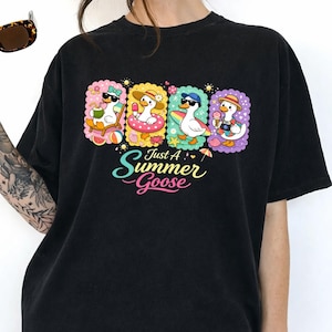 Pode incluir: Camiseta preta com um gráfico colorido de quatro gansos de desenho animado aproveitando as atividades de verão. Cada ganso está em uma cena de verão diferente, com o texto "Just A Summer Goose" em uma fonte divertida abaixo.