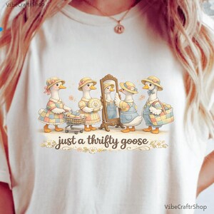 Pode incluir: Camiseta creme com uma ilustração fantasiosa de quatro gansos vestidos com roupas vintage. Os gansos estão a fazer compras e a olhar para um espelho. O texto "just a thrifty goose" está por baixo.