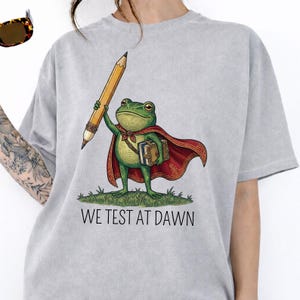 Puede incluir: Camiseta gris claro con un dibujo de una rana de dibujos animados vestida de superhéroe, sosteniendo un lápiz y libros. La rana lleva una capa roja y una mochila marrón. El texto "WE TEST AT DAWN" está impreso debajo.