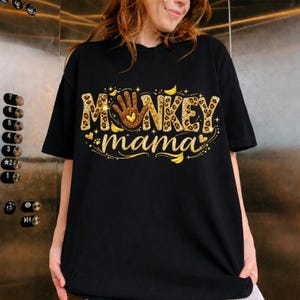 Può includere: T-shirt nera con le parole "MONKEY mama" in un design dorato e leopardato. La parola "MONKEY" presenta una mano con un cuore al centro. Il design include cuori dorati e illustrazioni di banane.