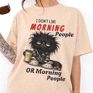 Op de afbeelding: Beige t-shirt met een chagrijnige zwarte kat in een badjas en konijnenslippers, met een koffiekopje. De tekst luidt: "I DON'T LIKE MORNING People OR Morning People."