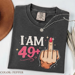 May include: Dark gray t-shirt with the text "I AM 49+" and a graphic of a hand giving the middle finger. The shirt has a crew neck and short sleeves. A silver necklace is draped across the shirt.