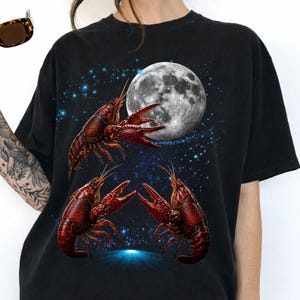 May include: Black t-shirt featuring a graphic design of three red crawfish in front of a full moon and a starry night sky. The crawfish are detailed with visible claws and antennae. The design has a celestial theme.