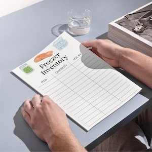 Puede incluir: Un bloc de notas blanco para el inventario del congelador con las palabras "Freezer Inventory" y secciones para artículo, cantidad y fechas de caducidad. El bloc de notas tiene ilustraciones de alimentos. Un vaso de agua está cerca.