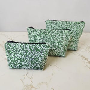 May include: Three green zippered pouches with a white swirl pattern. The pouches are in varying sizes, with black zippers. The fabric has a repeating design of white swirls on a green background. The pouches are arranged on a marble surface.