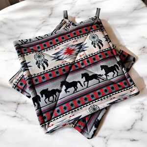 May include: Two square pot holders with a Native American design. The gray fabric features black horse silhouettes, dream catchers, and geometric patterns in red, black, and turquoise. Each pot holder has a gray loop for hanging.