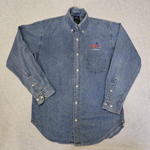 Peut inclure: Chemise en jean bleu clair à manches longues. La chemise a une petite poche sur la poitrine et un logo sur le côté gauche. Elle est dotée de boutons blancs et d'un col boutonné.