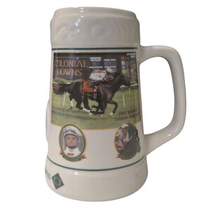 May include: Cream-coloured ceramic beer stein with a racing scene featuring a jockey on a black horse. The stein has a handle and a decorative top. The text "COLONIAL DOWNS" is visible, along with "2007 Winner Summer Derby".