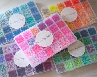May include: A collection of colorful craft supplies, including multiple clear plastic organizers filled with small, flat, round beads in various shades. The organizers are arranged to showcase a spectrum of colors, from blues and greens to pinks and oranges. Each organizer has a circular label that reads "Rave Grey Designs".