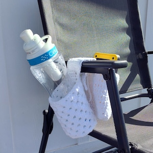 以下が含まれることがあります： 黒い金属製の椅子に、白い編み物のカップホルダーが掛けられています。ホルダーの中には水筒が入っています。椅子は黒いメッシュ素材で、黄色のプラスチック製のハンドルが付いています。