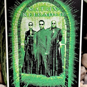 May include: A black and green poster with the title "MATRIX RELOADED" in white text. Three figures in black clothing stand in a green doorway. The figures are surrounded by a green and white digital effect.