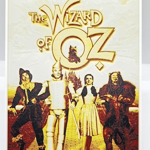 May include: A framed print featuring the characters from The Wizard of Oz. The image includes the Scarecrow, Tin Man, Dorothy, and the Cowardly Lion, with the title text in gold. The background is a yellow brick road.