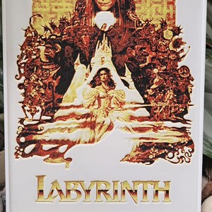 May include: A framed print featuring the movie "Labyrinth." The artwork showcases characters in shades of red, gold, and black against a maze-like background. The title "LABYRINTH" is displayed in gold lettering at the bottom.
