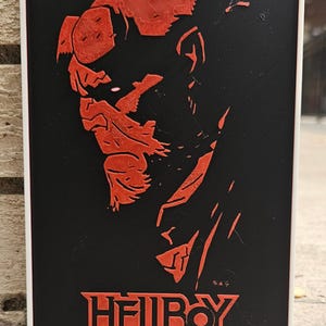 May include: A black and red Hellboy art print. The image features a profile of Hellboy in red against a black background. The word "HELLBOY" is printed in red at the bottom. The print is framed in a white border.