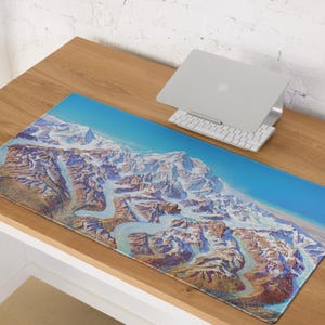 May include: A rectangular desk mat featuring a detailed illustration of a mountain range with snow-capped peaks and winding glacial rivers. The mat has a blue sky gradient and is placed on a wooden desk, next to a laptop and keyboard.