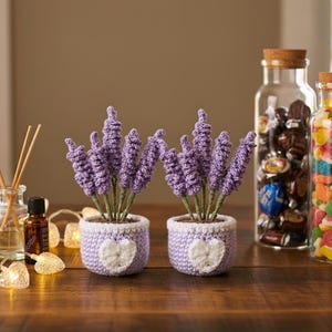 May include: Two crochet lavender plants in small, light purple pots with white heart designs. The lavender has purple blooms and green stems. The pots sit on a wooden surface with decorative jars and lights in the background.