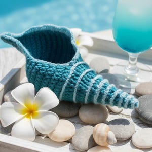 May include: A turquoise crochet seashell-shaped container with white spiral accents. The container sits on a white tray with smooth stones, a white flower, and a blue drink in a glass. The seashell is a decorative item.
