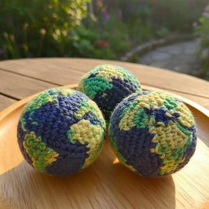 May include: Three crocheted Earth globes in a wooden tray. The globes feature blue oceans, green continents, and yellow landmasses. The handmade spheres are displayed in natural sunlight, showcasing the texture of the yarn.