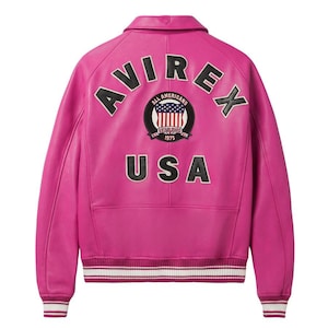 K&ouml;nnte beinhalten: Eine leuchtend pinkfarbene Lederjacke mit dem Wort "AVIREX" &uuml;ber den Schultern. Auf dem R&uuml;cken befindet sich ein kreisf&ouml;rmiger Aufn&auml;her mit den Worten "ALL AMERICANS ESTABLISHED 1975" und einem USA-Flaggen-Design. Das Wort "USA" ist darunter gedruckt. Die Jacke hat einen gerippten Saum und B&uuml;ndchen.