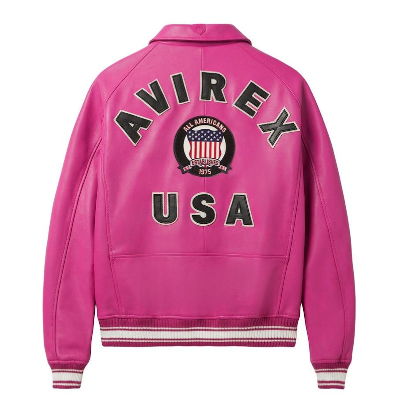 K&ouml;nnte beinhalten: Eine leuchtend pinkfarbene Lederjacke mit dem Wort "AVIREX" &uuml;ber den Schultern. Auf dem R&uuml;cken befindet sich ein kreisf&ouml;rmiger Aufn&auml;her mit den Worten "ALL AMERICANS ESTABLISHED 1975" und einem USA-Flaggen-Design. Das Wort "USA" ist darunter gedruckt. Die Jacke hat einen gerippten Saum und B&uuml;ndchen.
