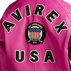 K&ouml;nnte beinhalten: Nahaufnahme der R&uuml;ckseite einer leuchtend rosa Lederjacke. Die Jacke hat schwarz-wei&szlig;e gestickte Buchstaben mit der Aufschrift "AVIREX" und "USA". Ein kreisf&ouml;rmiger Aufn&auml;her mit den Worten "ALL AMERICANS" und "ESTABLISHED 1975" ist ebenfalls zu sehen.