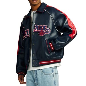 Puede incluir: Chaqueta de cuero azul marino con detalles rojos y blancos. La chaqueta tiene cierre de botones, bolsillos laterales y cuello blanco. Las mangas tienen rayas rojas y la chaqueta tiene la palabra "ACE" en cursiva.