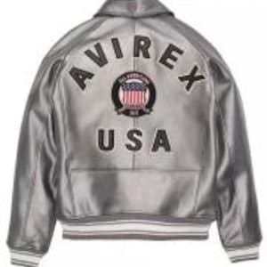 Puede incluir: Chaqueta bomber plateada con el texto "AVIREX USA" bordado en la espalda. La chaqueta presenta un dise&ntilde;o cl&aacute;sico con cuello, pu&ntilde;os y dobladillo de canal&eacute;. La chaqueta tiene un brillo met&aacute;lico y un parche con la bandera americana.