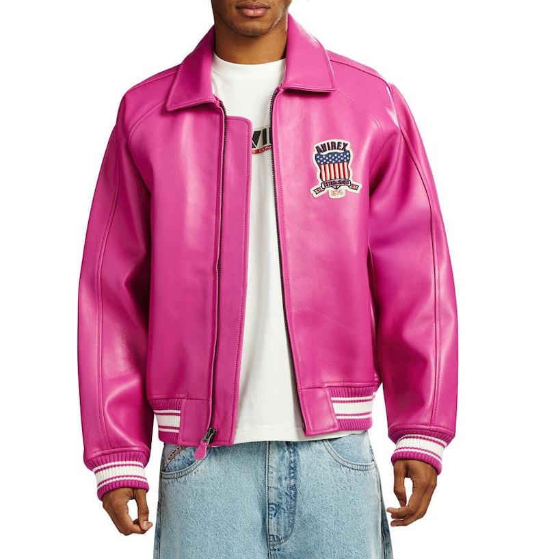 K&ouml;nnte beinhalten: Eine leuchtend pinkfarbene Lederjacke im klassischen Bomberjacken-Design. Mit Kragen, Rei&szlig;verschluss vorne und gerippten B&uuml;ndchen und Saum mit wei&szlig;en Streifen. Ein gestickter Aufn&auml;her mit patriotischem Design auf der Brust. Ein auff&auml;lliges Kleidungsst&uuml;ck.