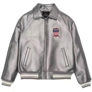 Puede incluir: Chaqueta de cuero plateada con cremallera, cuello y pu&ntilde;os acanalados con rayas blancas. Cuenta con un parche en el pecho con un dise&ntilde;o de bandera americana y el nombre de la marca. Un estilo cl&aacute;sico de chaqueta bomber.