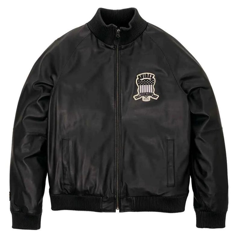 May include: Black leather jacket with a ribbed collar, cuffs, and hem. Features a full-length zipper and a patch on the chest with the text "AVIREX" and a shield design. A classic bomber jacket style.