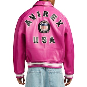 K&ouml;nnte beinhalten: Eine leuchtend pinkfarbene Lederjacke mit dem Wort "AVIREX" in schwarzen Buchstaben auf dem R&uuml;cken. Die Jacke hat einen Aufn&auml;her mit "ALL AMERICANS" und "USA" darunter. Die Jacke hat einen wei&szlig;-schwarzen Streifen am &Auml;rmel und am Bund.