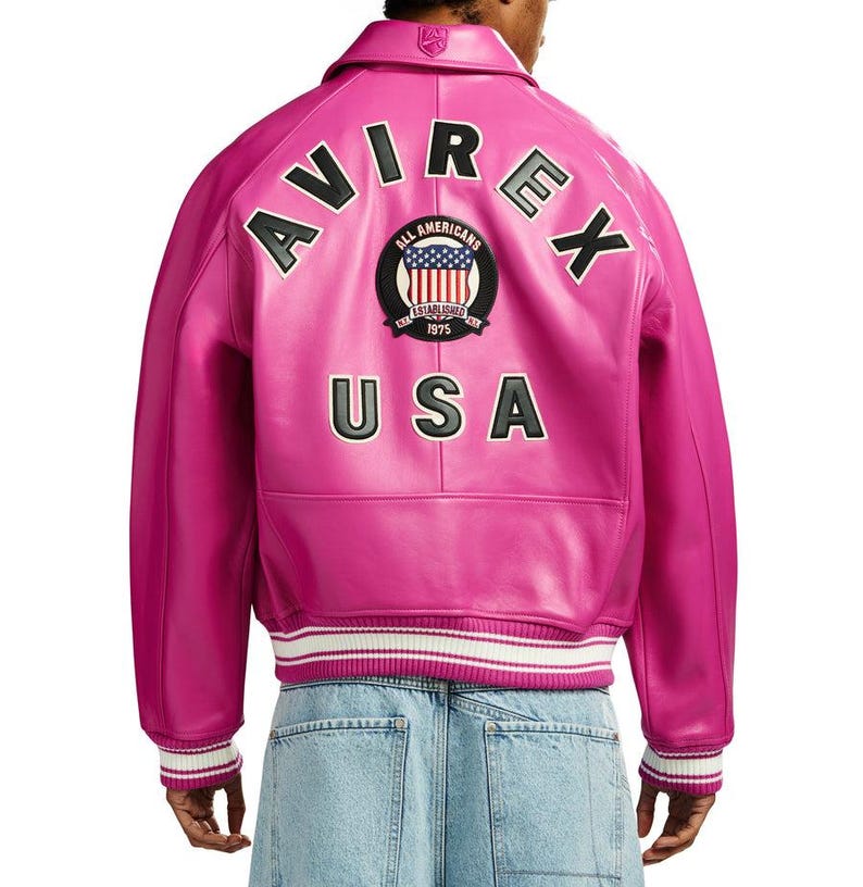 K&ouml;nnte beinhalten: Eine leuchtend pinkfarbene Lederjacke mit dem Wort "AVIREX" in schwarzen Buchstaben auf dem R&uuml;cken. Die Jacke hat einen Aufn&auml;her mit "ALL AMERICANS" und "USA" darunter. Die Jacke hat einen wei&szlig;-schwarzen Streifen am &Auml;rmel und am Bund.