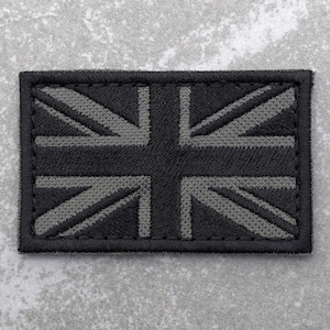 May include: A black and grey embroidered patch featuring the Union Jack flag. The patch has a black border and is made of a woven fabric.